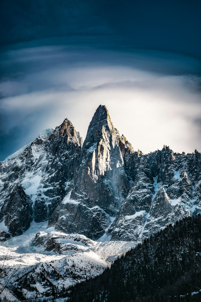 A breathtaking late afternoon scene in the Chamonix Valley, Haute-Savoie, where mountain peaks pierce the sky, gracefully enveloped by a solitary cloud, illuminated by the gently setting sun, creating a magnificent display of contrasts and volumes against a backdrop of a deeply-hued sky, with a tranquil, dark coniferous forest adorning the foreground.
