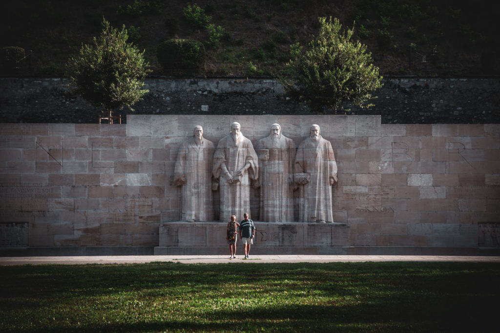 Lumière envoûtante baignant la statue de Calvin au Mur des Réformateurs avec visiteurs se dirigeant vers l'ombre