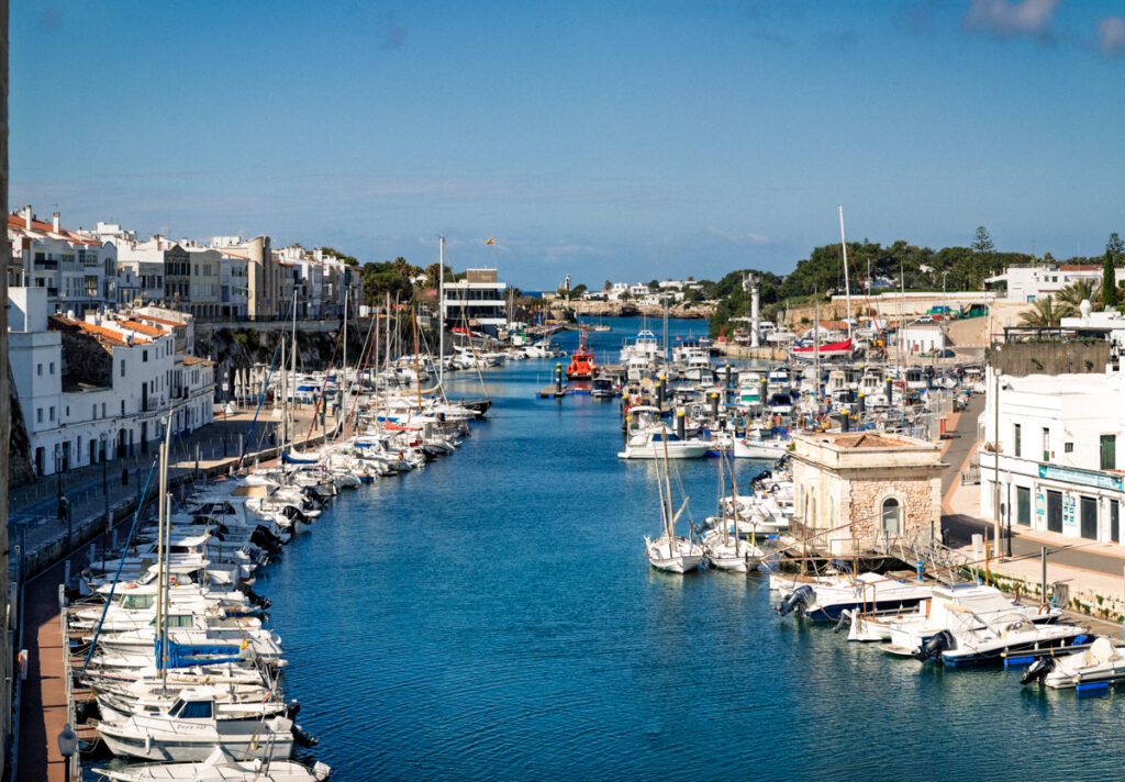 Vue plongeante sur le vieux port de Ciutadella, chenal d'eau bleue bordé de bateaux de pêche et de voiliers entre les façades blanches, ouverture vers la mer au fond, Minorque