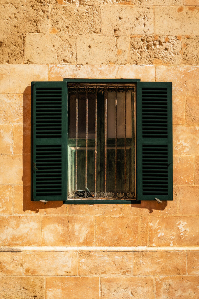 Fenêtre aux volets persiennés verts et barreaux de fer forgé dans un mur de calcaire doré, Ciutadella, Minorque