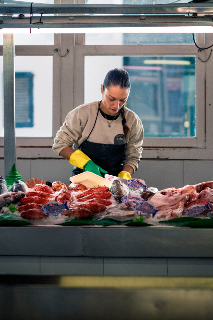 Une poissonnière en tablier prépare le poisson derrière un étal de rougets et sargues sur glace, marché couvert de Ciutadella, Minorque