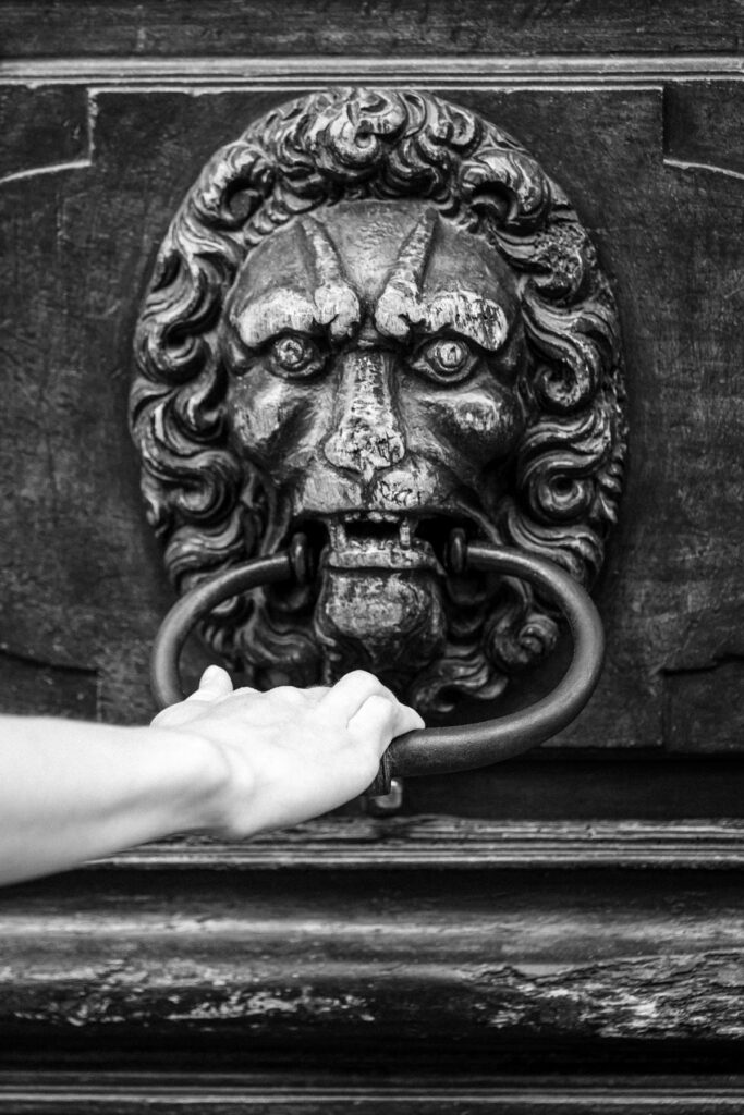 De Passage — main saisissant l'anneau de bronze d'un heurtoir en tête de lion, Vieille-Ville de Genève, noir et blanc
